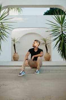 A stylish young man sits casually in a modern outdoor space surrounded by greenery.